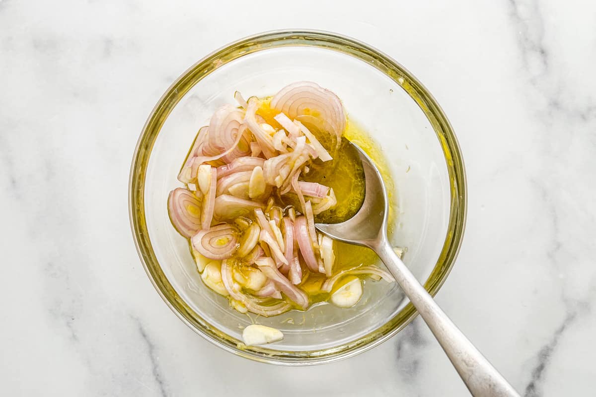 A small glass bowl with olive oil, lemon juice, sliced garlic, and sliced shallots.