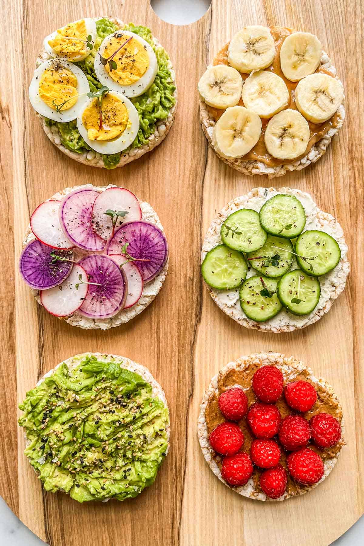 Six rice cakes with healthy toppings on a wooden cutting board.