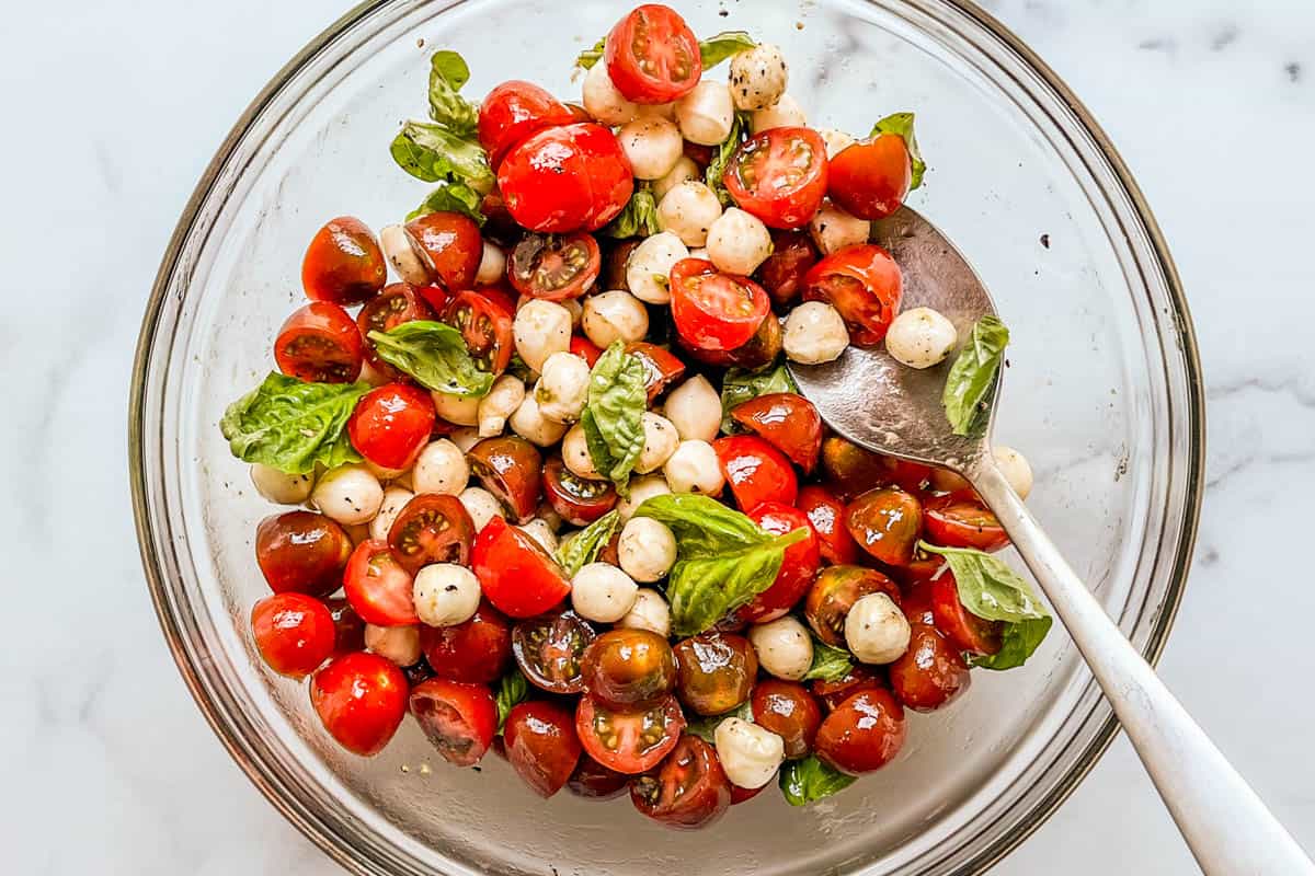 A cherry tomato caprese salad being tossed in a glass bowl.