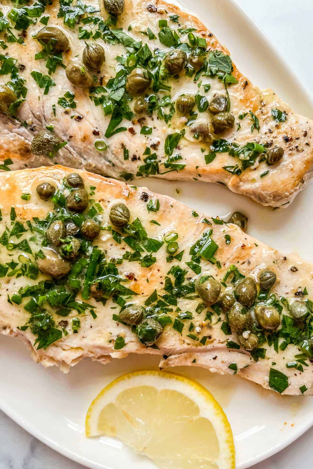 Two swordfish steaks with a herb butter caper sauce on a white platter.