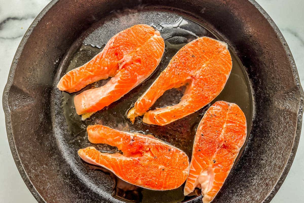 Four salmon steaks being cooked in a cast iron pan.
