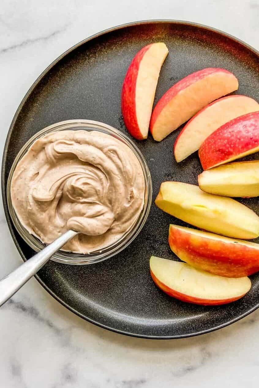 A small bowl of Greek yogurt dessert dip next to apple slices on a black plate.