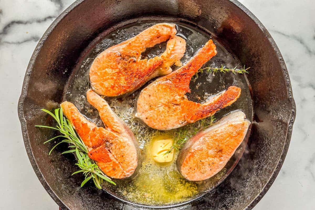 Salmon steaks being cooked in butter and herbs in a cast iron pan.
