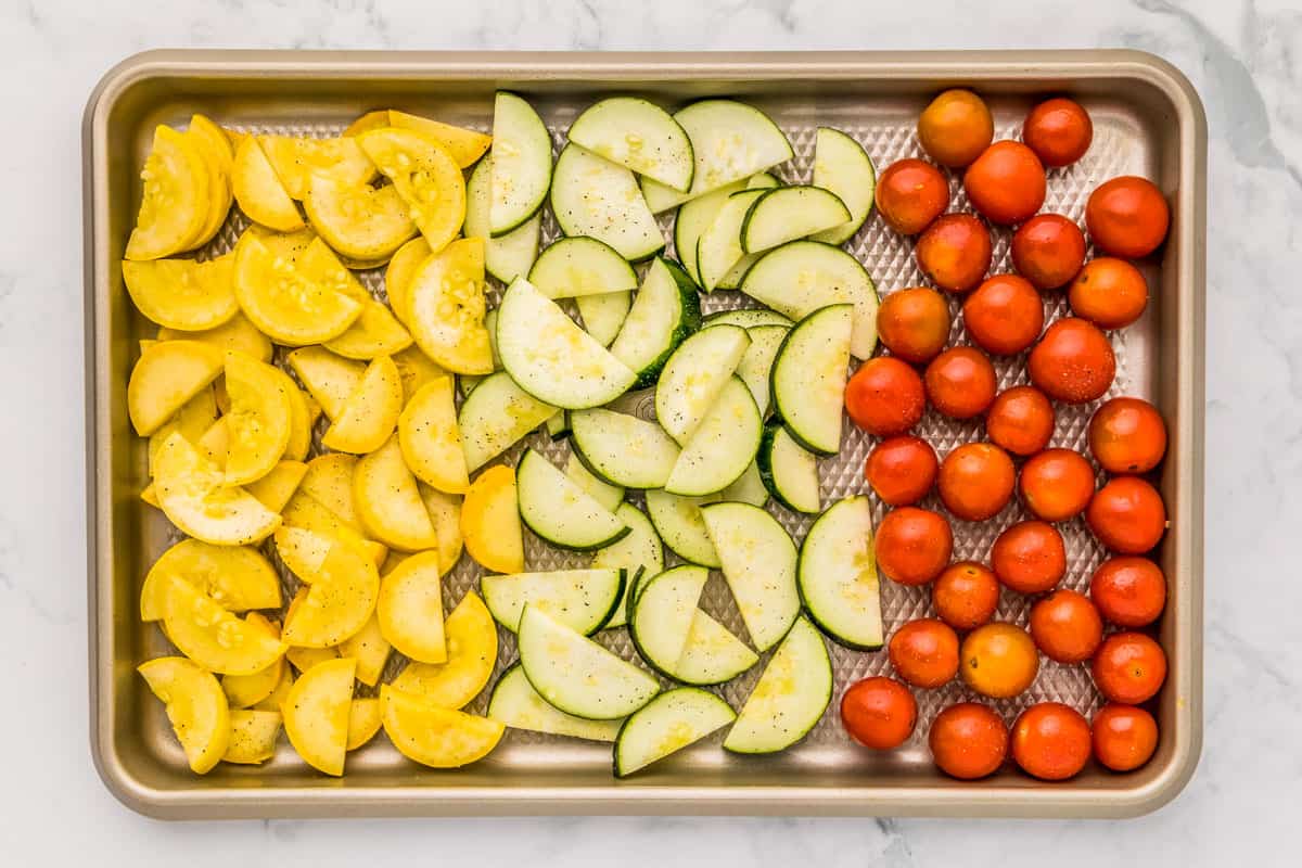 Sliced yellow squash, zucchini, and cherry tomatoes on a rimmed baking sheet.