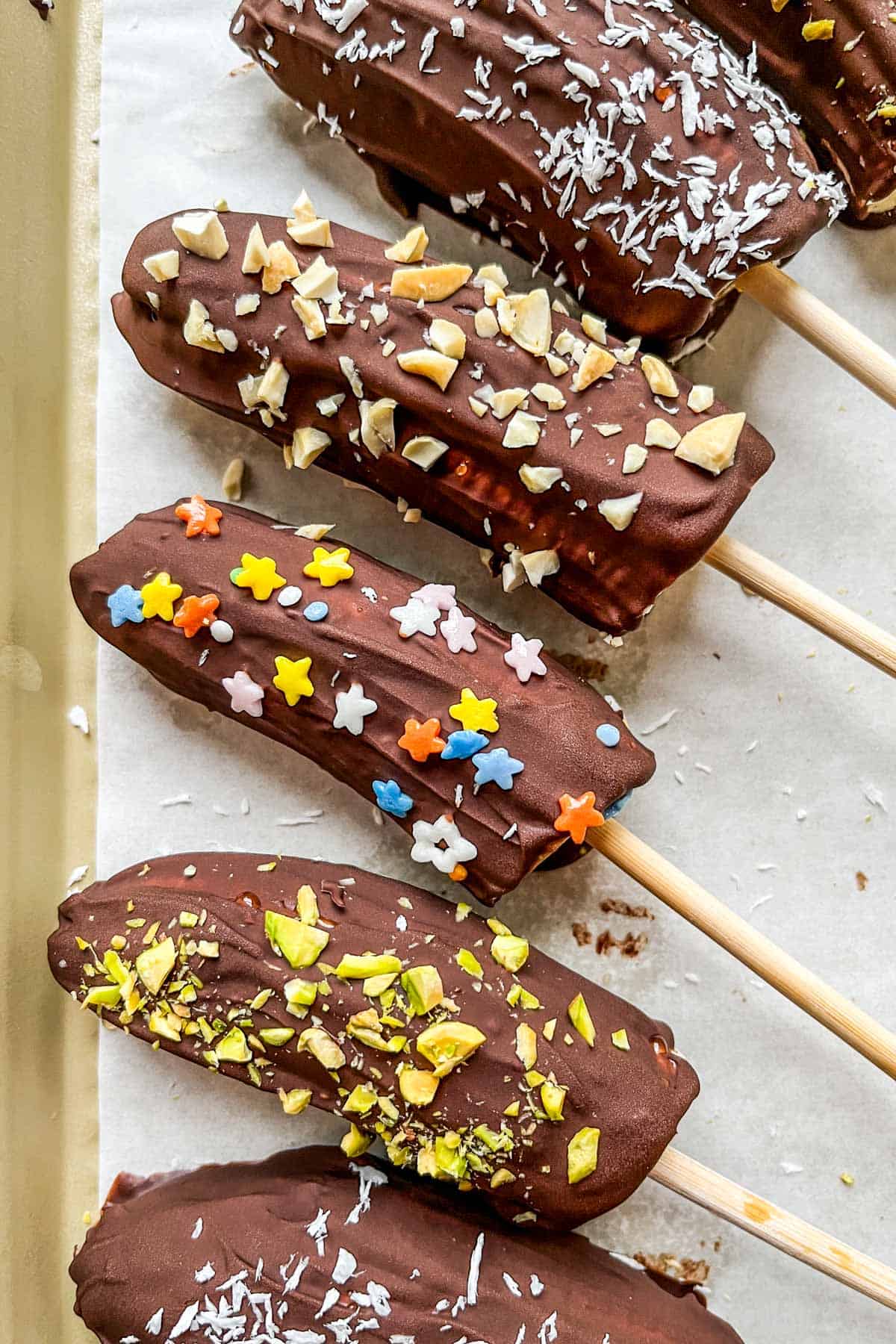 Dark chocolate bananas decorated with sprinkles, chopped nuts, and coconut flakes on a parchment lined baking sheet.
