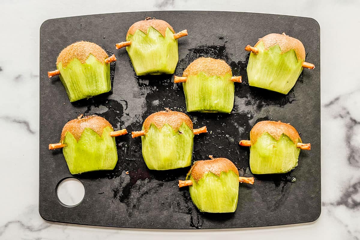 Kiwis with pretzels sticks on the edges on a cutting board.
