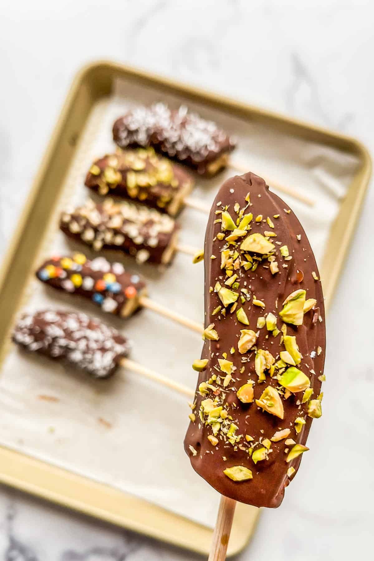 A chocolate covered banana with pistachios being held in front of a baking sheet with more chocolate bananas.