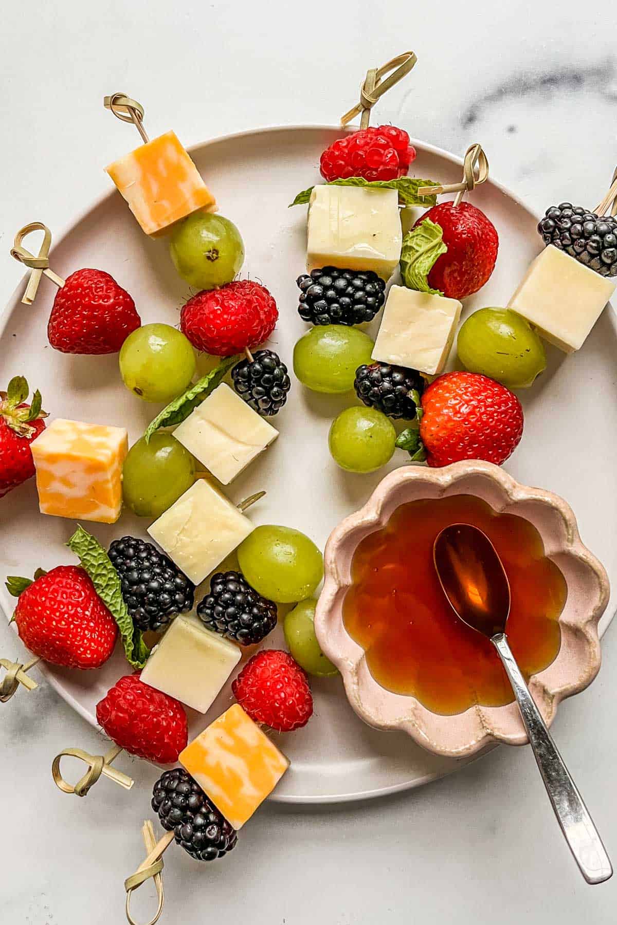 Fruit and cheese skewers on a white plate with a small bowl of honey.