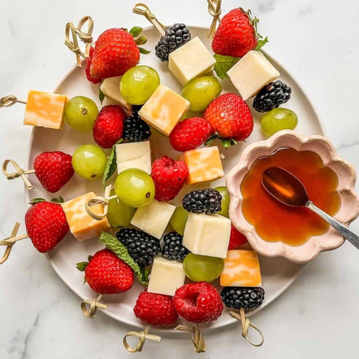 Fruit and cheese kabobs on a white plate with a small bowl of honey.