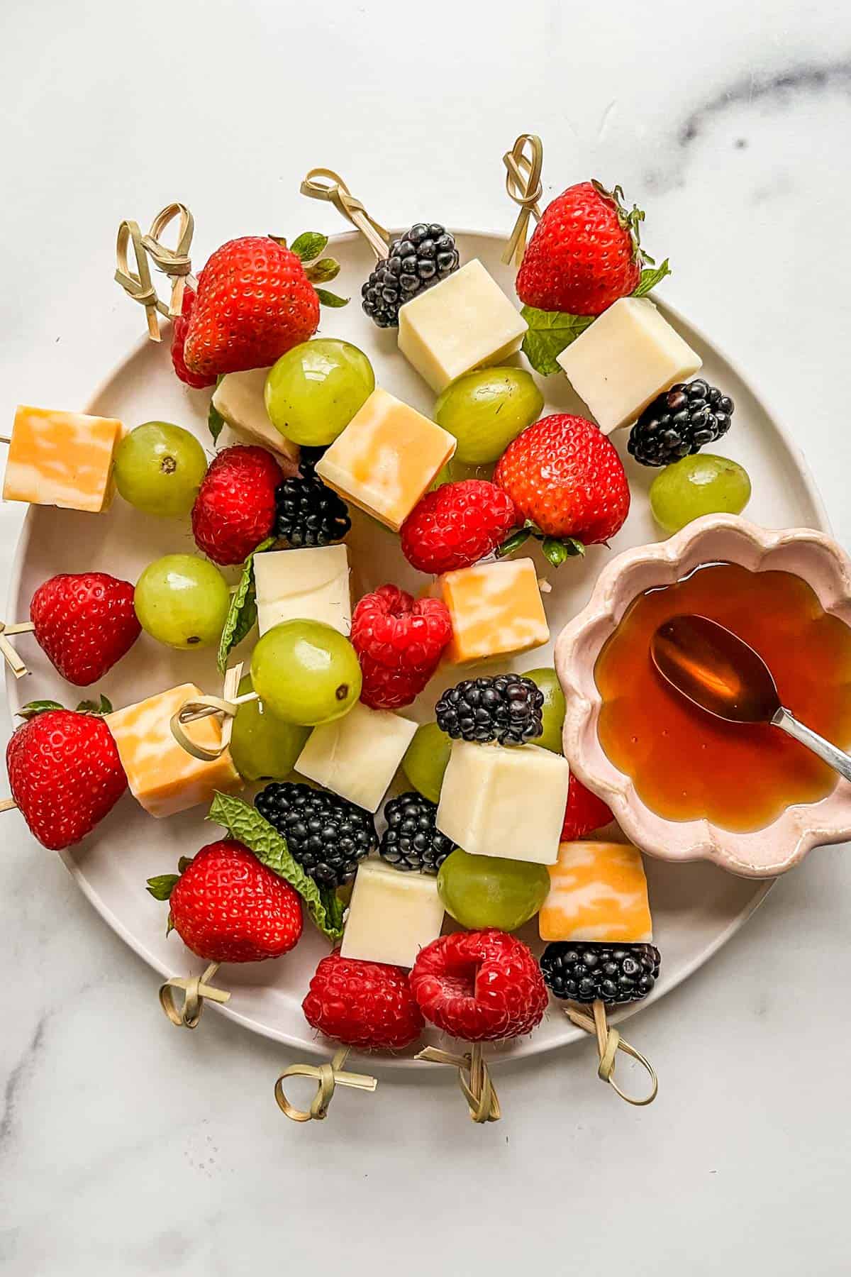 Fruit and cheese skewers on a white plate with a small bowl of honey.