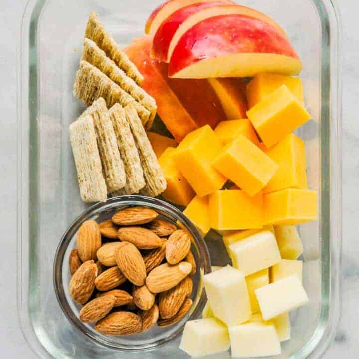An adult lunchable with cheese, crackers, apple slices, and almonds in a glass container.