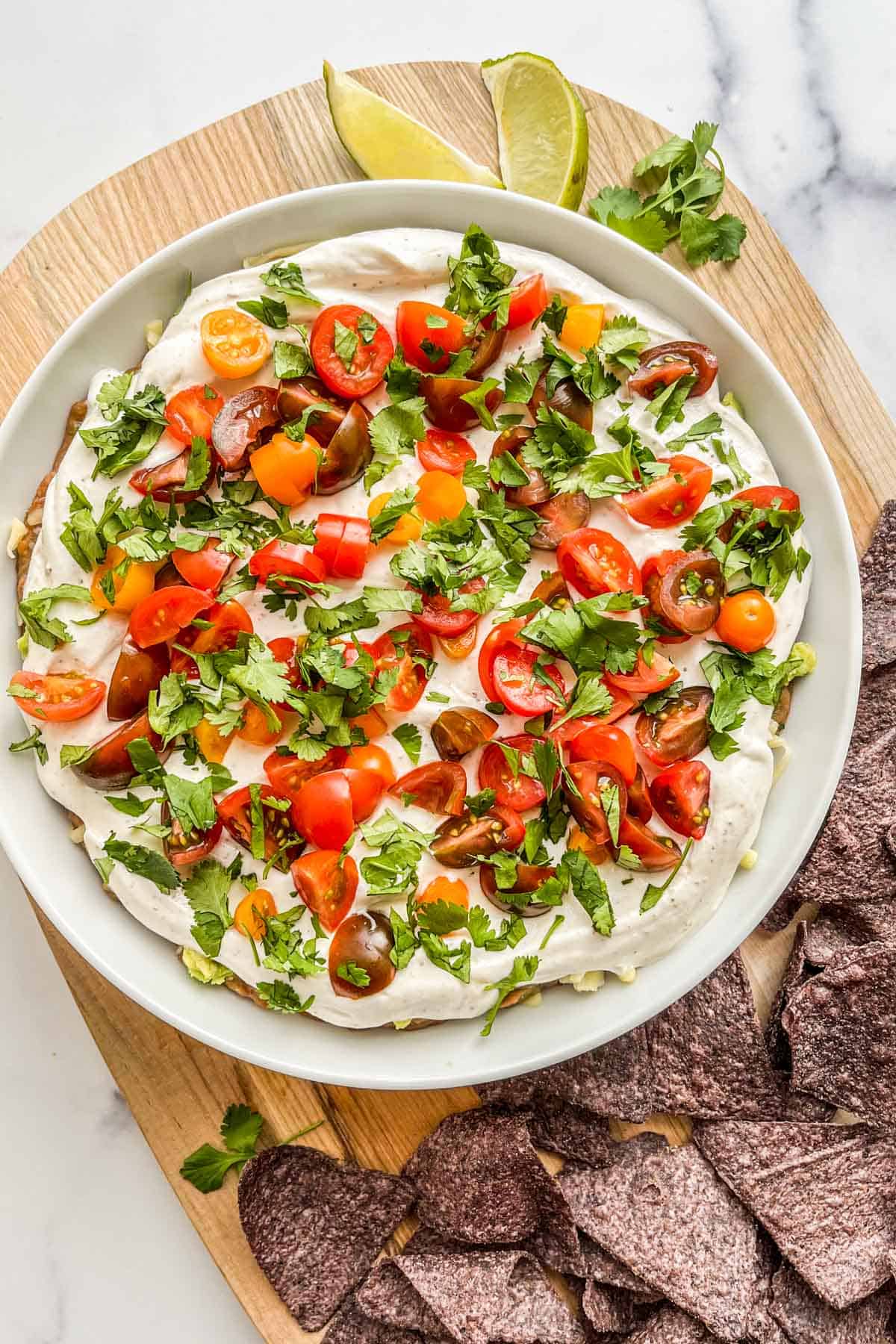 A 7-layer dip in a large white bowl on a wooden board with blue corn chips.