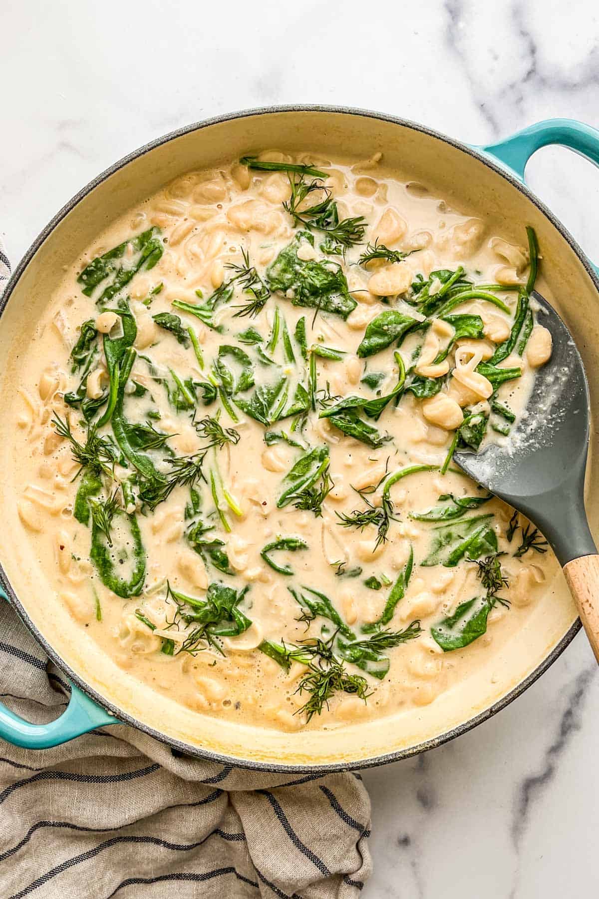 Creamy miso and spinach butter beans in a large pan with a spatula next to a striped towel.
