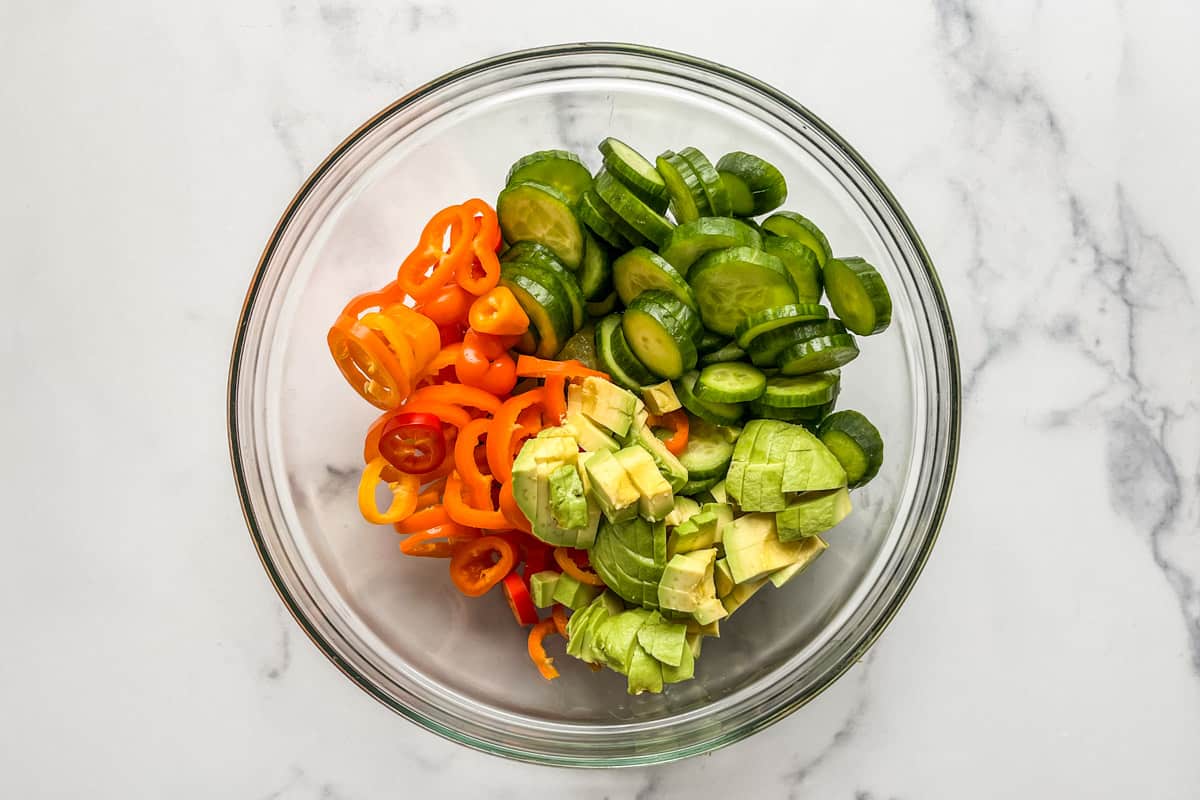 Sliced cucumbers, sweet peppers, and avocado in glass bowl.