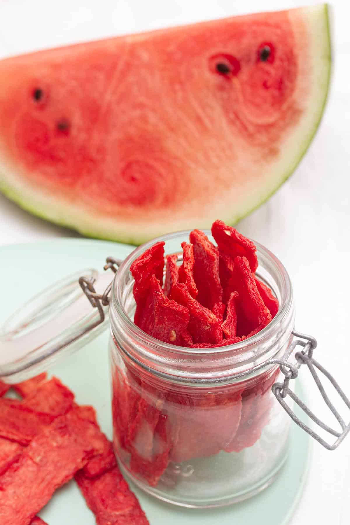 A jar with dehydrated watermelon, sitting in front of a slice of watermelon.