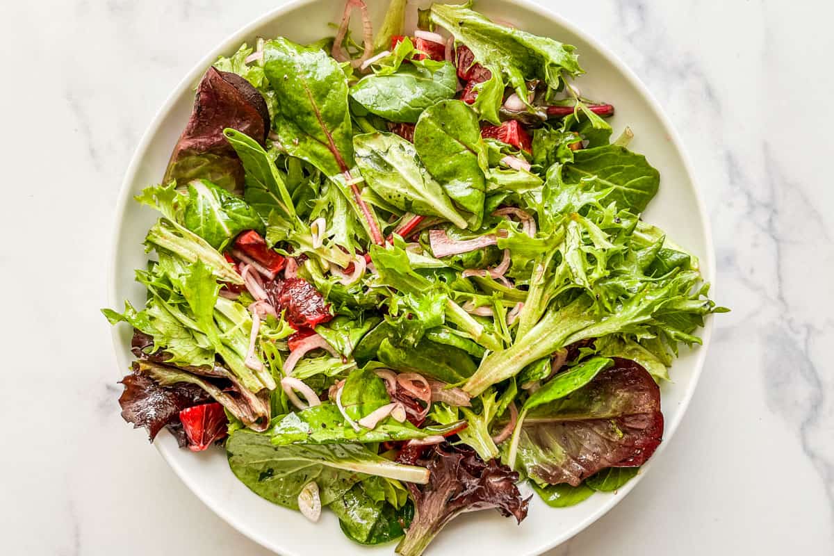 Greens, tossed with blood oranges, and sliced shallots in a white serving bowl.