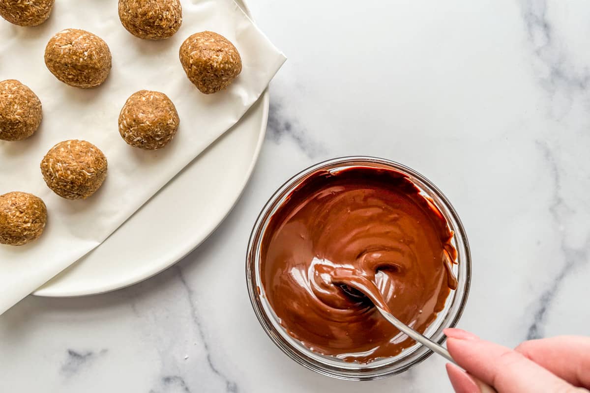 A small bowl of melted chocolate being stirred next to a plate with frozen coconut almond butter balls.