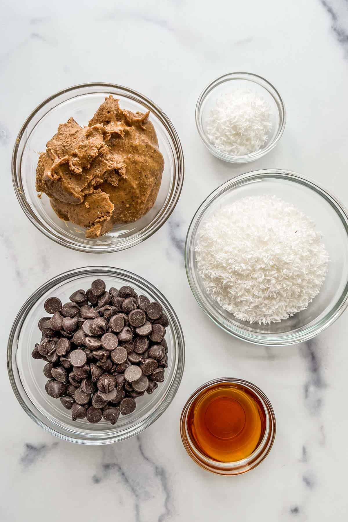 Coconut flakes, almond butter, dark chocolate chips, and maple syrup in glass bowls.