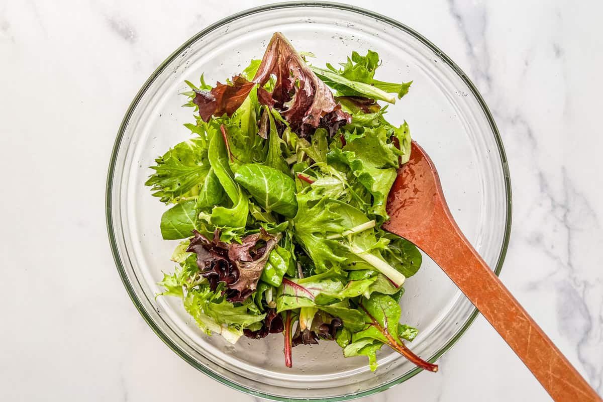 Greens tossed with a citrus honey salad dressing in a glass bowl with a wooden spoon.