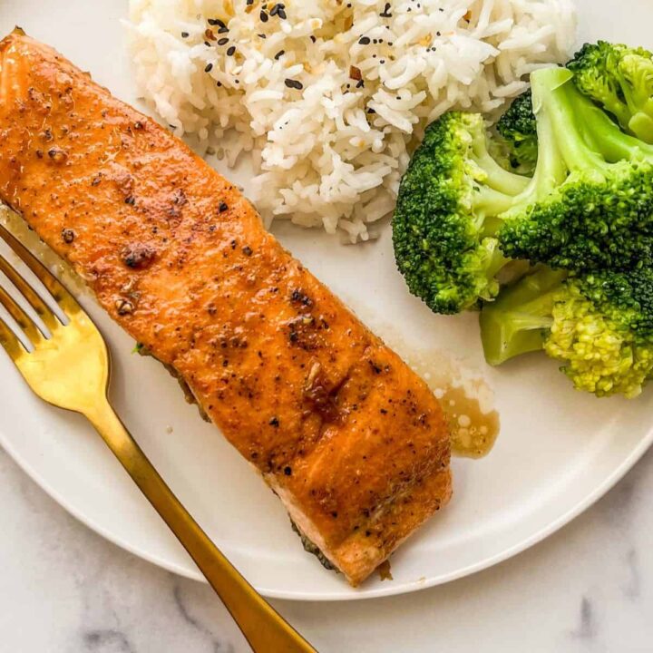 A maple glazed salmon fillet on a white plate with a gold fork next to steamed broccoli and white rice.