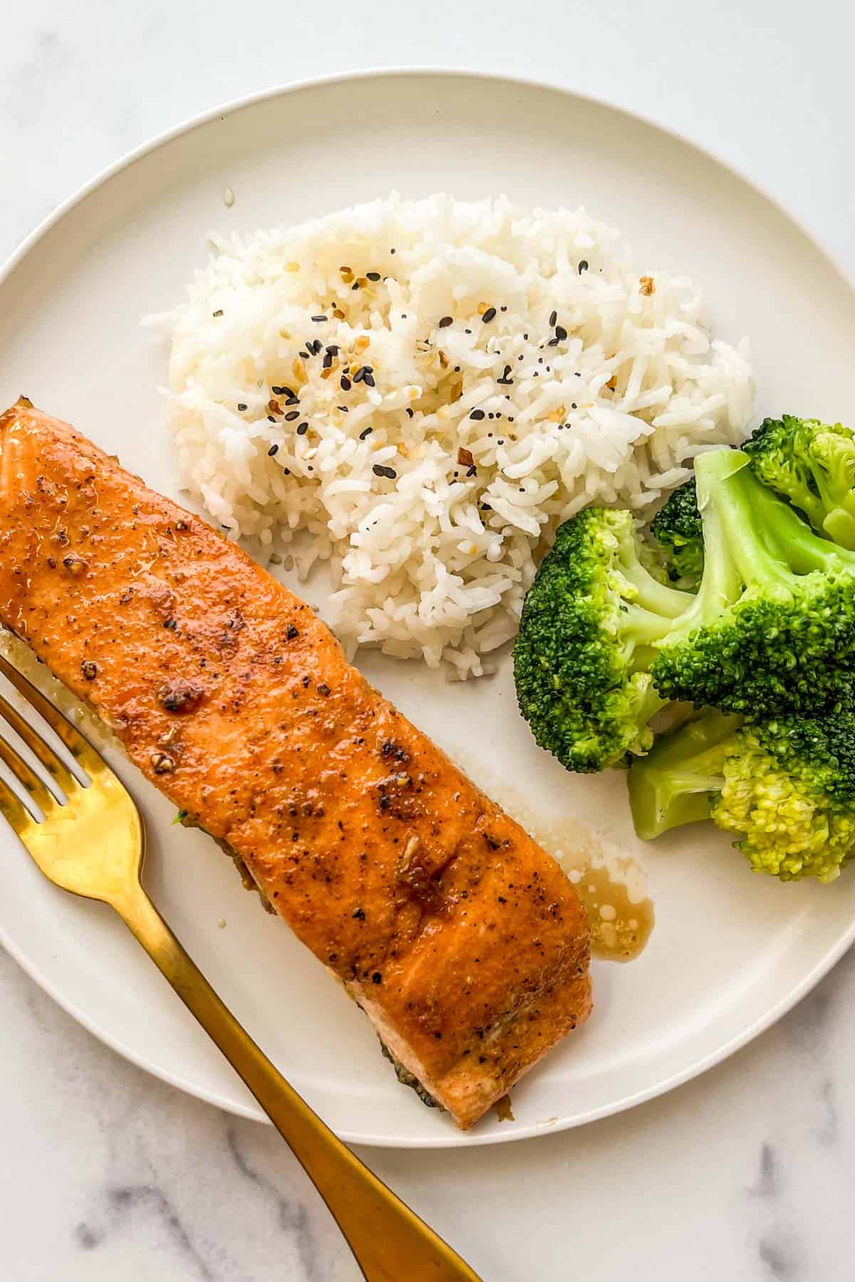 A maple glazed salmon fillet on a white plate with a gold fork next to steamed broccoli and white rice.