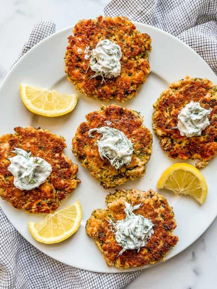 Salmon patties topped with dill sauce on a white plate with slices of lemon.