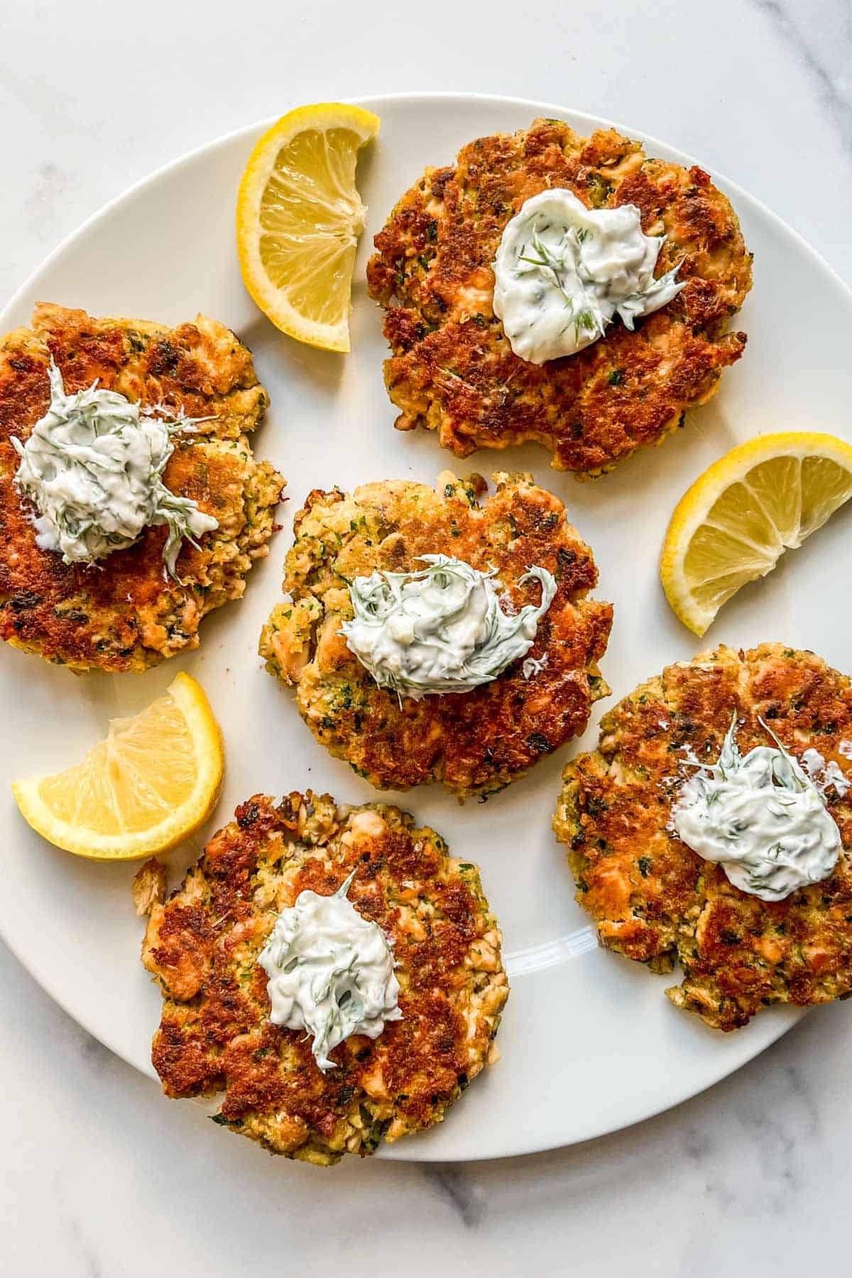 Salmon patties topped with dill sauce on a white plate with slices of lemon.