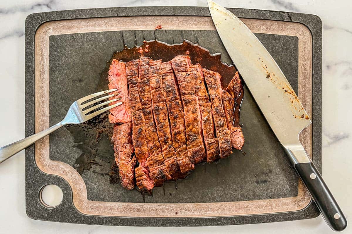 Flank steak cut into slices on a black cutting board with a fork and large knife.