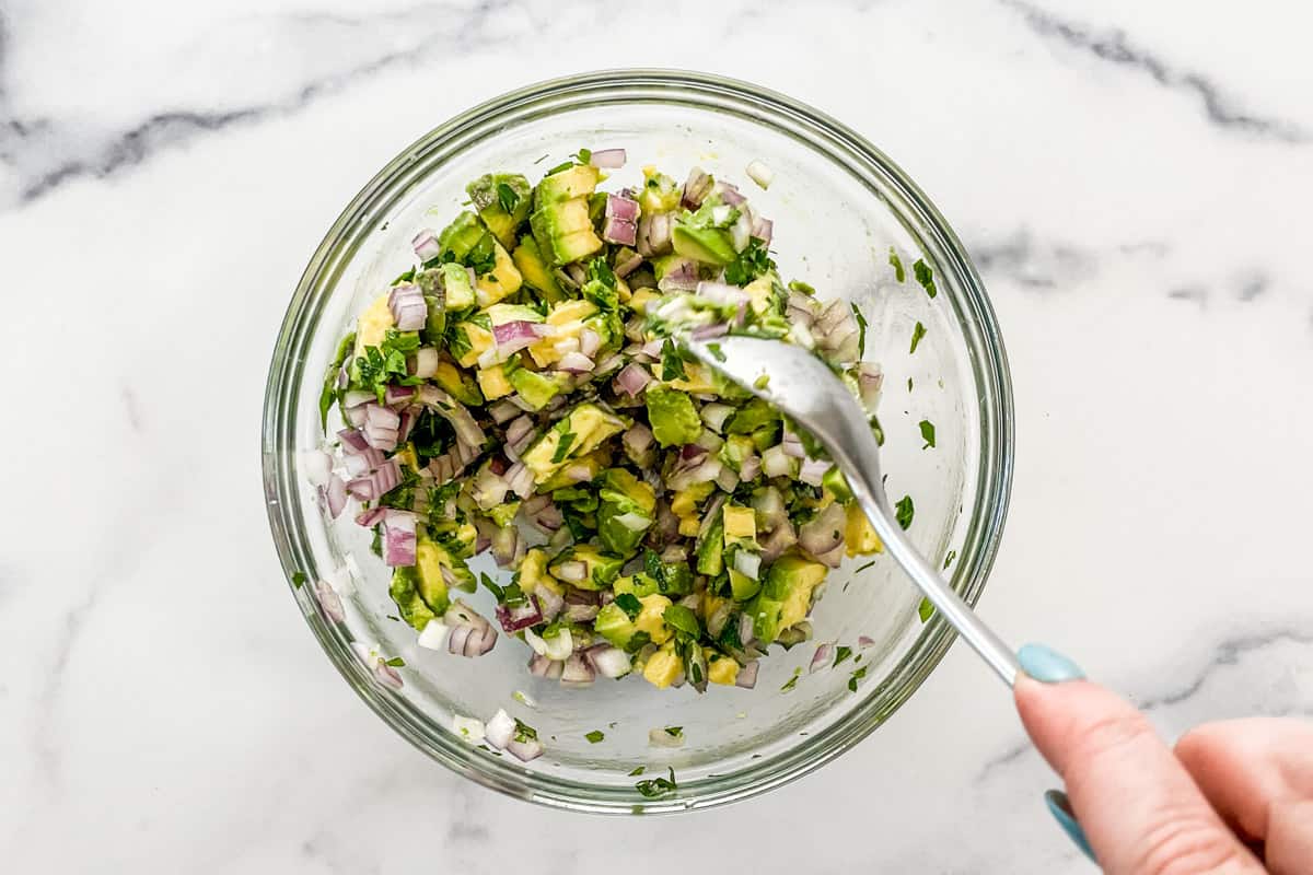 A glass bowl with a spoon stirring a mixture of chopped red onion, cilantro, and diced avocado.