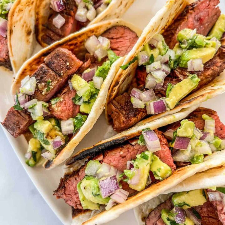 Flank steak tacos with chopped onion and avocado topping on a white plate.