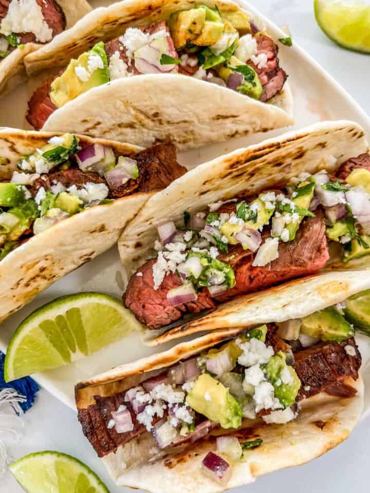 Grilled flank steak tacos on a white oval plate with lime wedges.