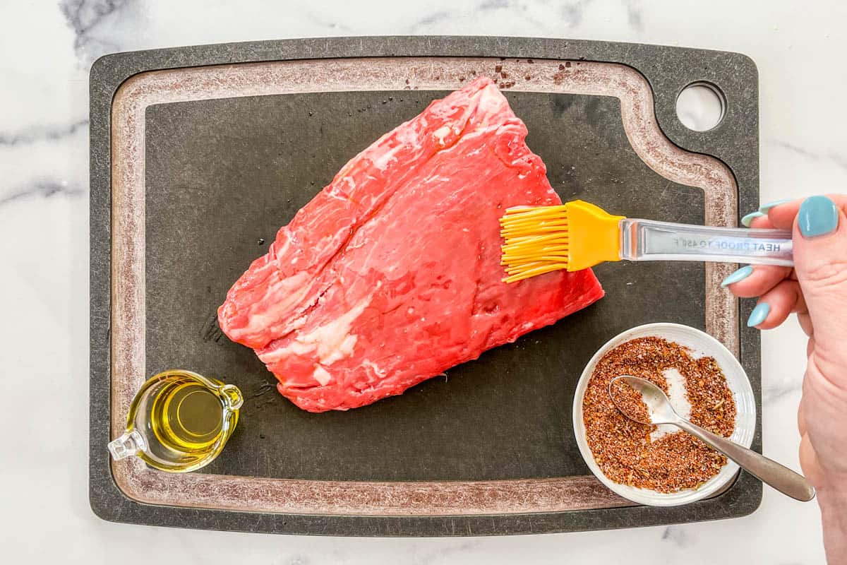 A silicone brush brushing olive oil onto a piece of flank steak on a black cutting board.