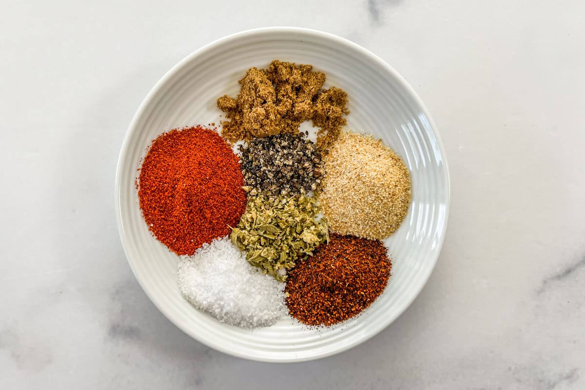 Paprika, cumin, garlic powder, chili powder, salt, dried oregano, and black pepper on a small white plate.