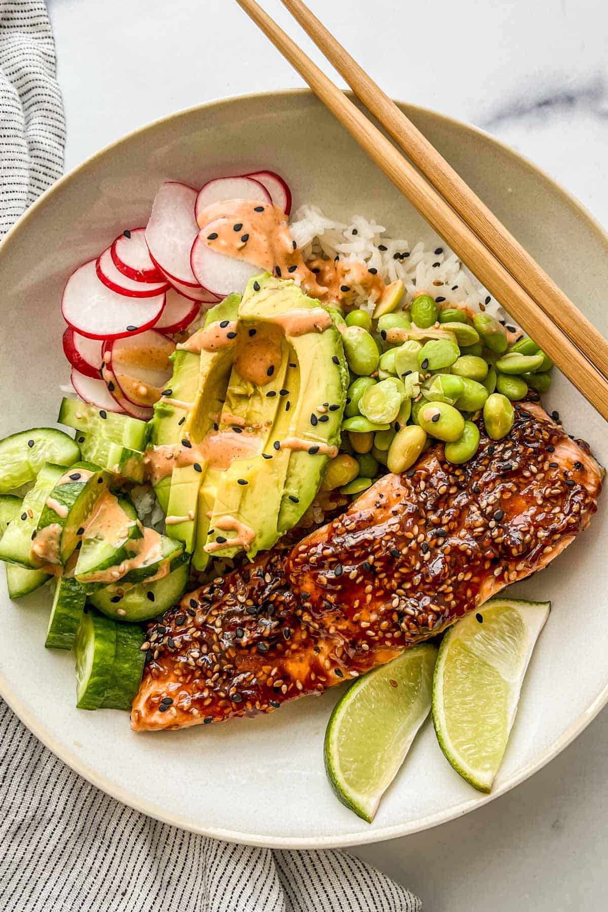 Teriyaki salmon fillet in a bowl with rice, avocado, cucumbers, edamame, radishes, and spicy mayo.