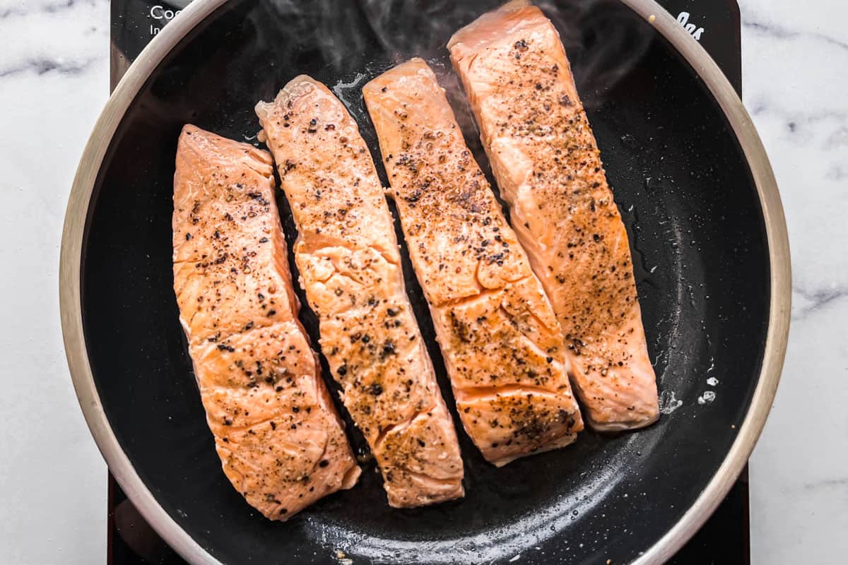 Four salmon fillets cooking in a frying pan.