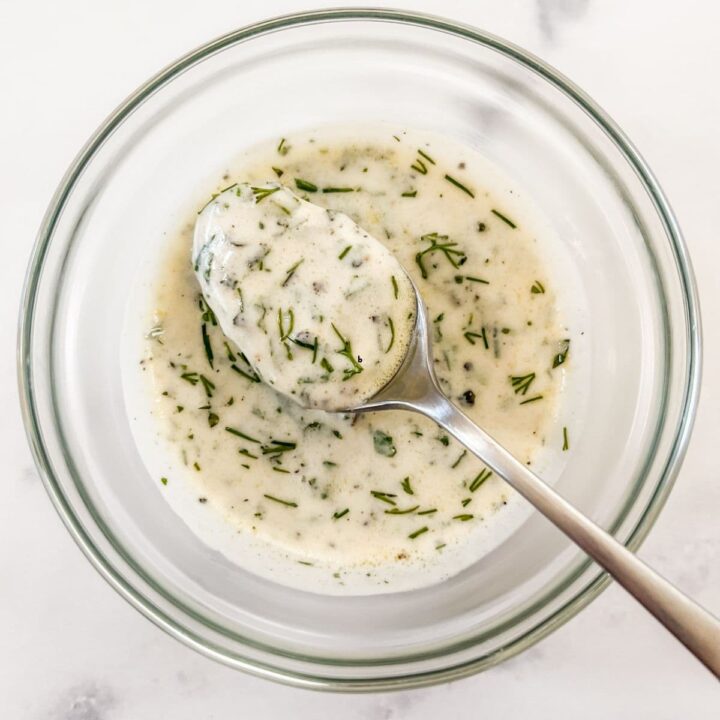 A silver spoon scooping up yogurt dill dressing from a glass bowl.