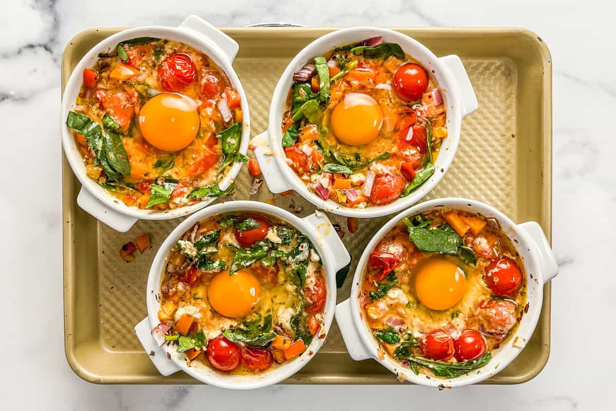 Four ramekins of feta, spinach, tomatoes, and raw eggs on a sheet pan.
