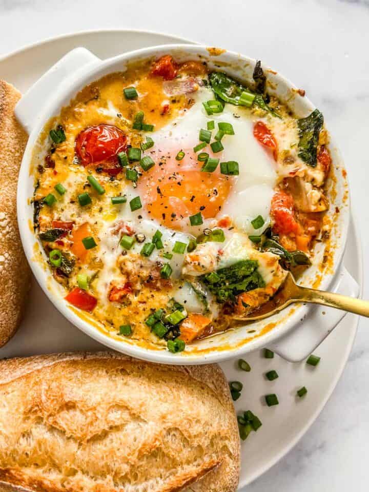 Baked feta eggs with spinach and tomatoes in a small ramekin with a gold spoon on a white plate with bread.
