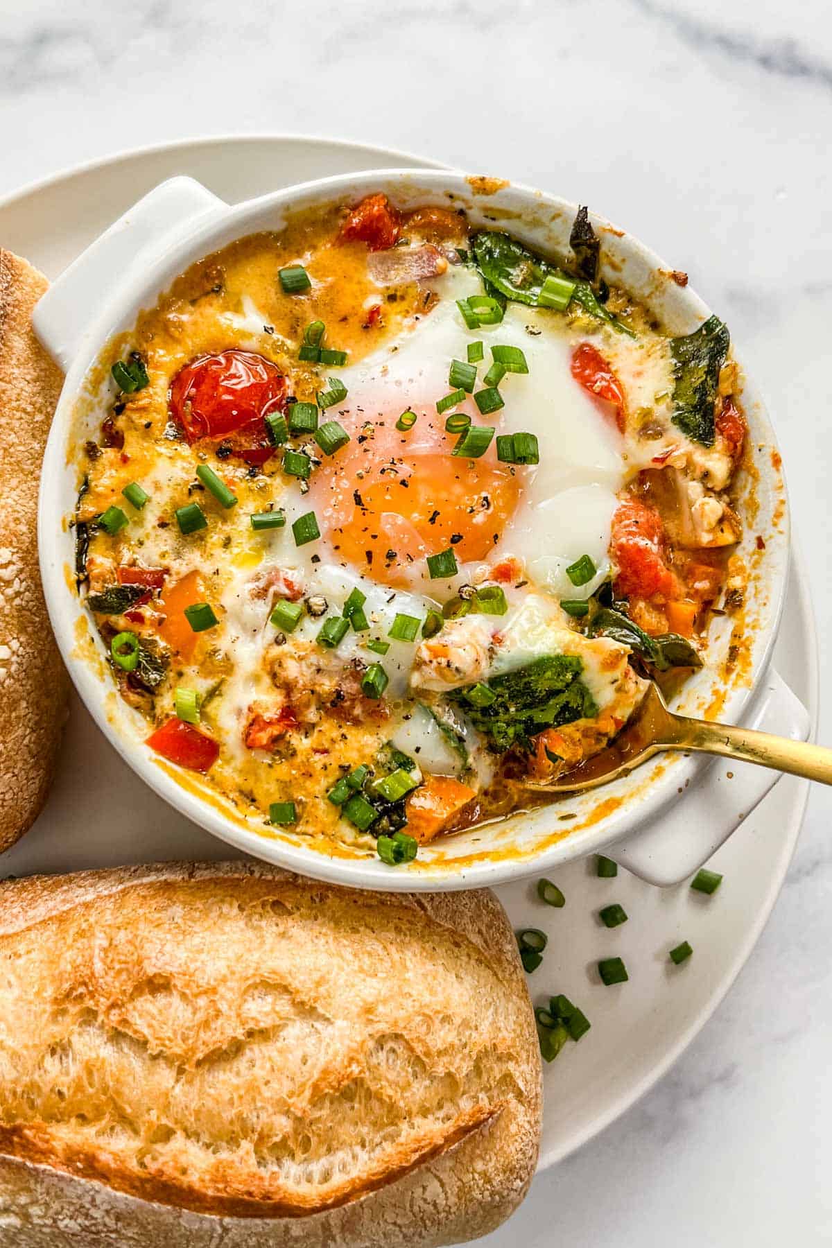 Baked feta eggs with spinach and tomatoes in a small ramekin with a gold spoon on a white plate with bread.