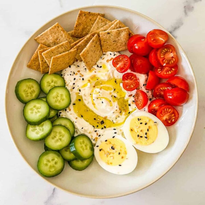 A savory yogurt bowl with crackers, cherry tomatoes, an egg, and sliced cucumbers.