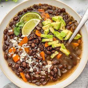 Black bean soup topped with limes, avocados, and feta cheese in an offwhite bowl with a silver spoon.
