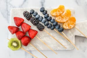 Wooden skewers with kiwi, strawberries, blackberries, blueberries, and oranges on a marble board.
