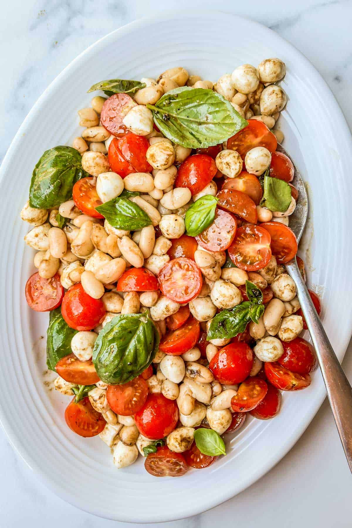 Caprese white bean salad with basil leaves on a white platter with a silver serving spoon.