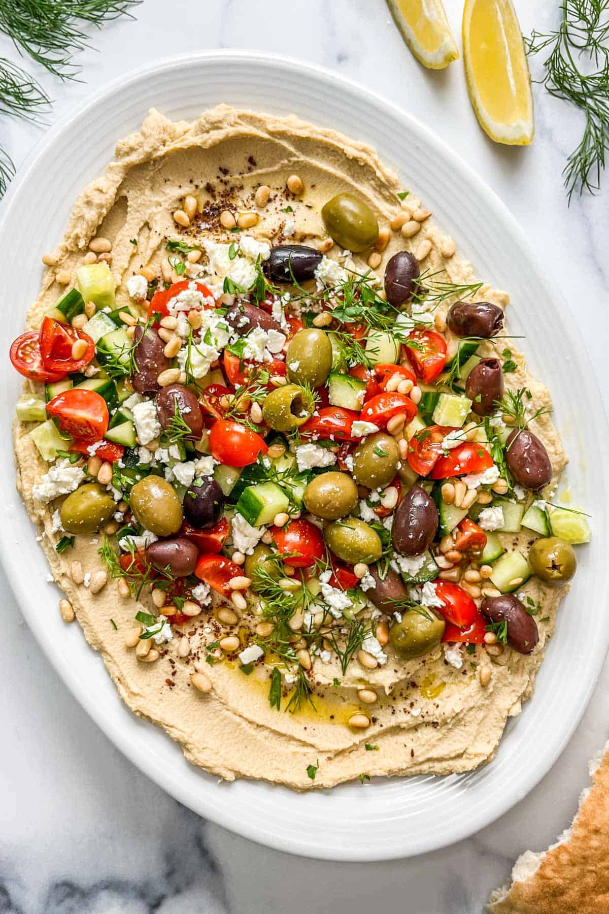 Loaded hummus with olives, tomatoes, cucumber, herbs, and toasted pine nuts on a white platter.