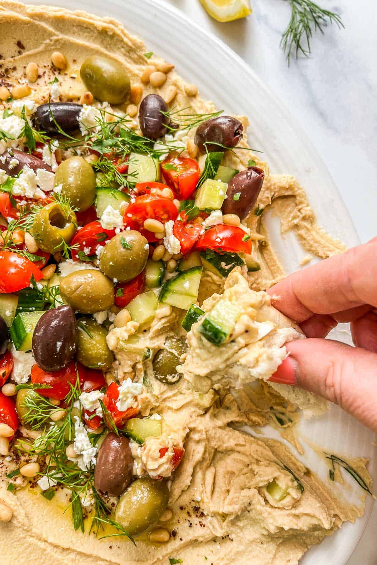 A hand scooping a piece of pita through loaded Greek hummus.