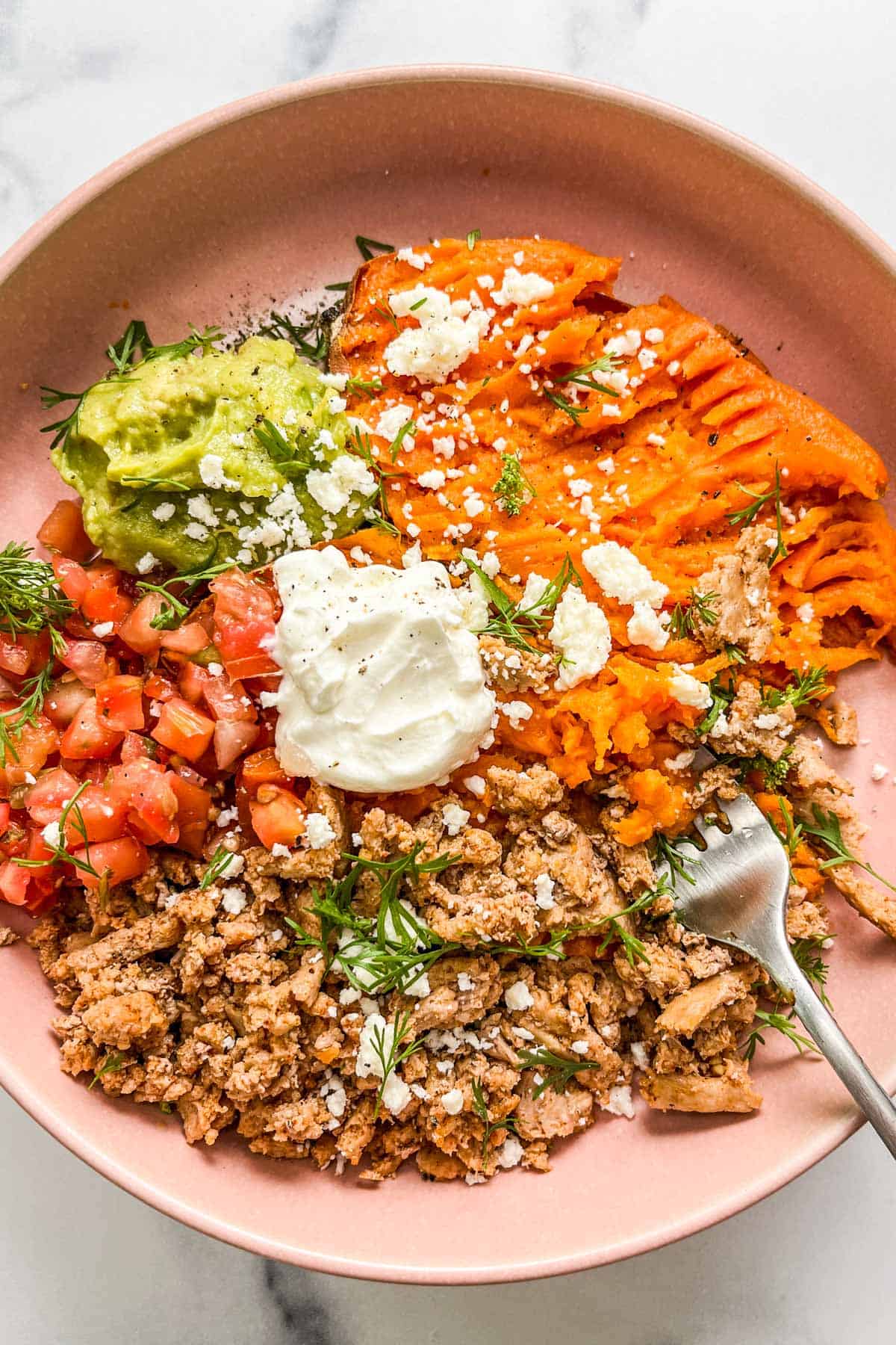A ground turkey sweet potato bowl with pico de gallo, guacamole, sour cream, crumbled cheese, herbs, and sliced lime in a pink bowl with a silver fork.