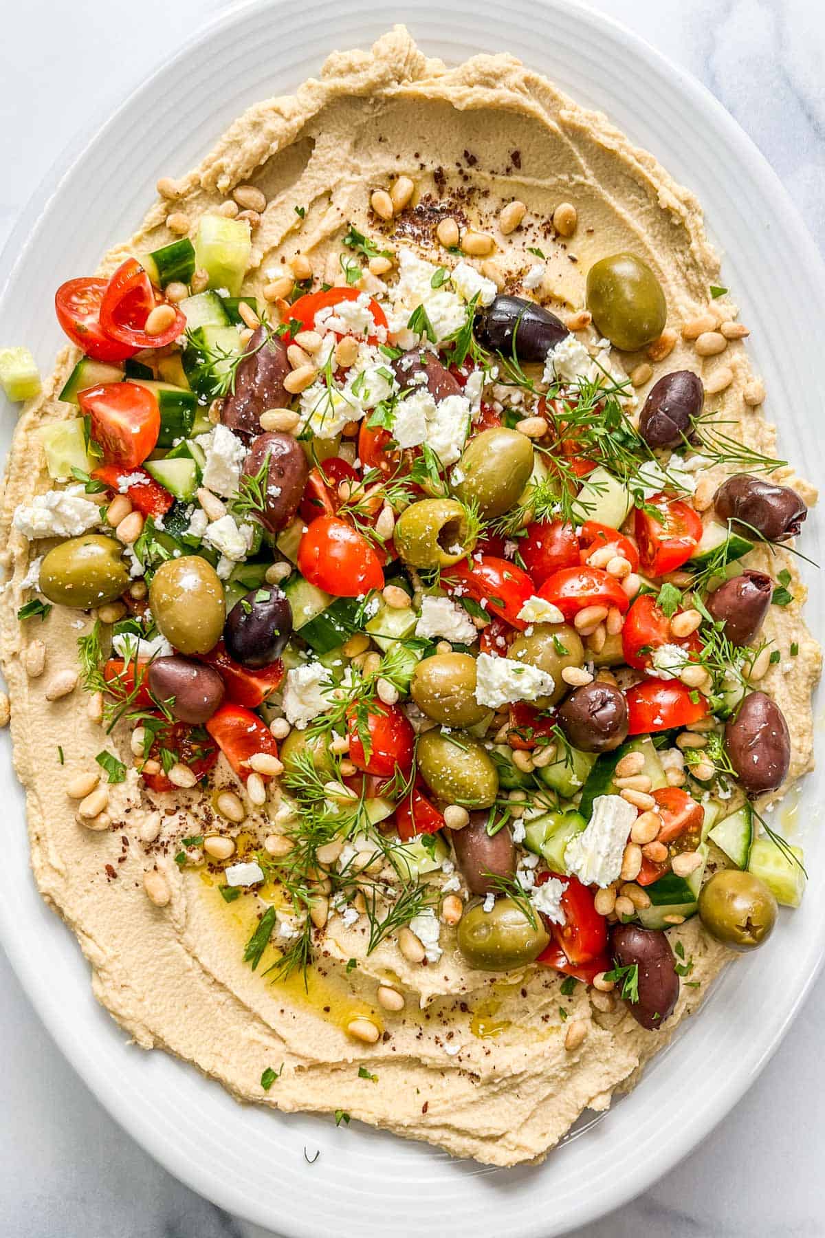Loaded hummus with olives, tomatoes, cucumber, herbs, and toasted pine nuts on a white platter.