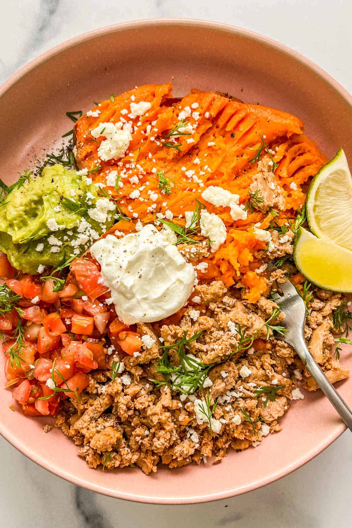 A ground turkey sweet potato bowl with pico de gallo, guacamole, sour cream, crumbled cheese, herbs, and sliced lime.
