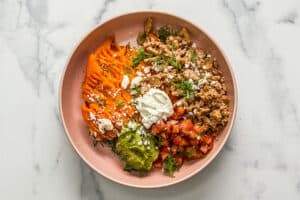 A ground turkey sweet potato bowl with pico de gallo, guacamole, sour cream, crumbled cheese, herbs, and sliced lime in a pink bowl.