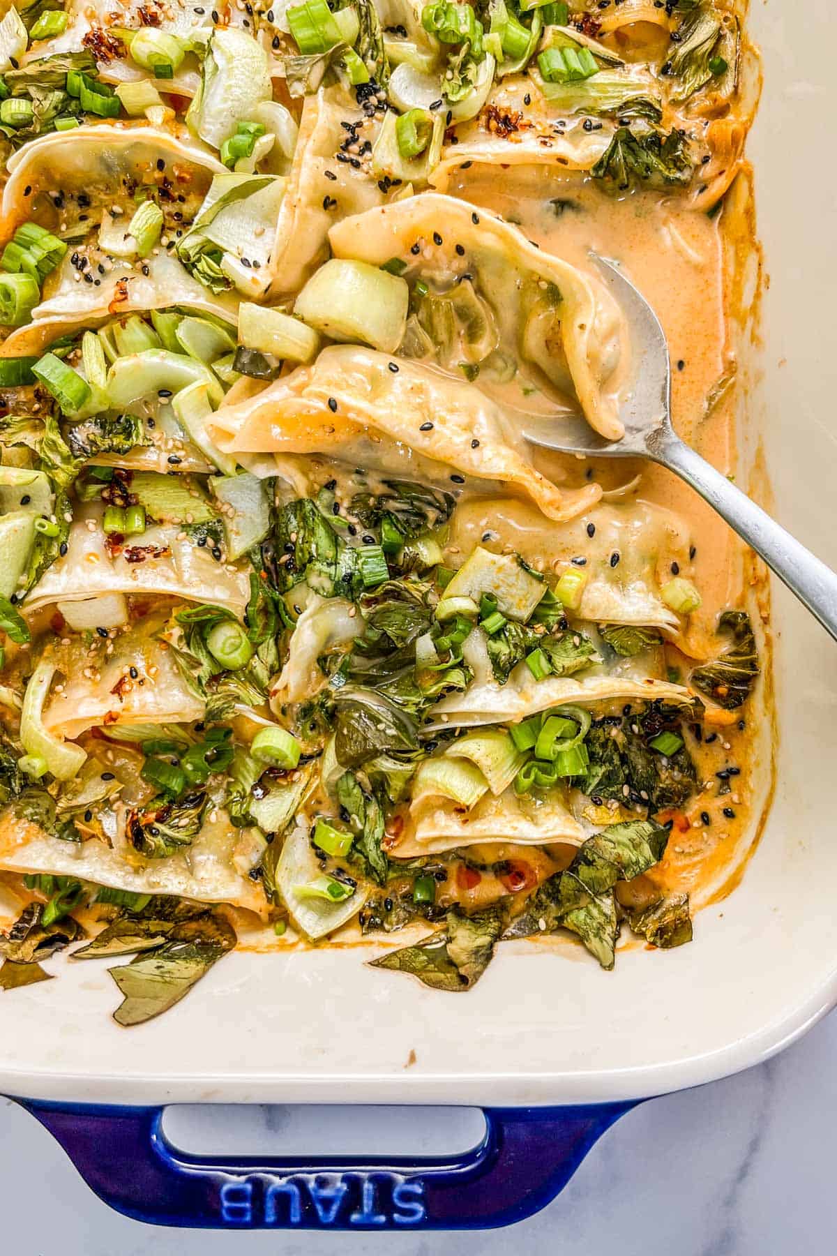 Casserole dish with baked dumplings topped with green onions, bok choy, sesame seeds, and chili oil and a silver spoon scooping them up.